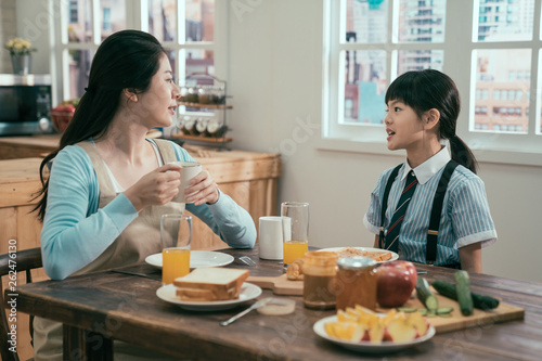 young asian korean mother and daughter having breakfast together in morning at home. mom in apron drinking hot coffee cup talking with cute little girl in school uniform. healthy meal on wooden table