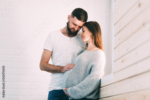 Young pregnant couple near the window of the house, on the background of a brick white wall and a wooden window sill © komokvm