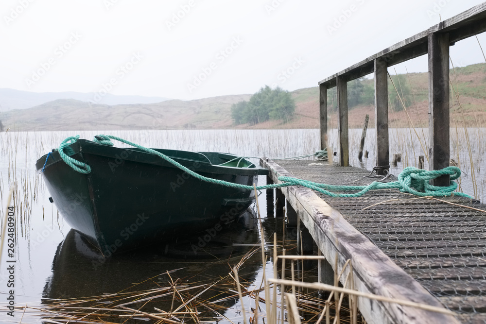 Fototapeta premium Zielona łódź wiosłowa przywiązana liną do drewnianego pomostu na jeziorze Loch w Argyll w Szkocji