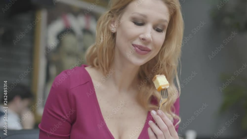 Attractive blond woman with curly hair eating cake with a macaroon in the restaurant close up. Hot lady with deep cleavage enjoys her dessert. Camera moves up Attractive blond woman with curly hair eating cake with a macaroon in the restaurant close up. Hot lady with deep cleavage enjoys her dessert. Camera moves up
