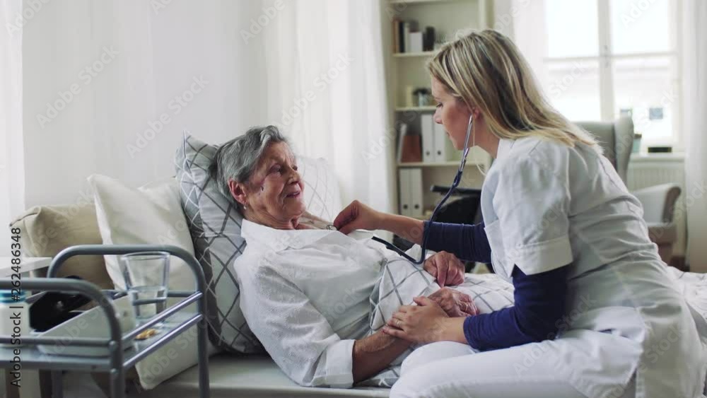 A health visitor examining a sick senior woman lying in bed at home with stethoscope.