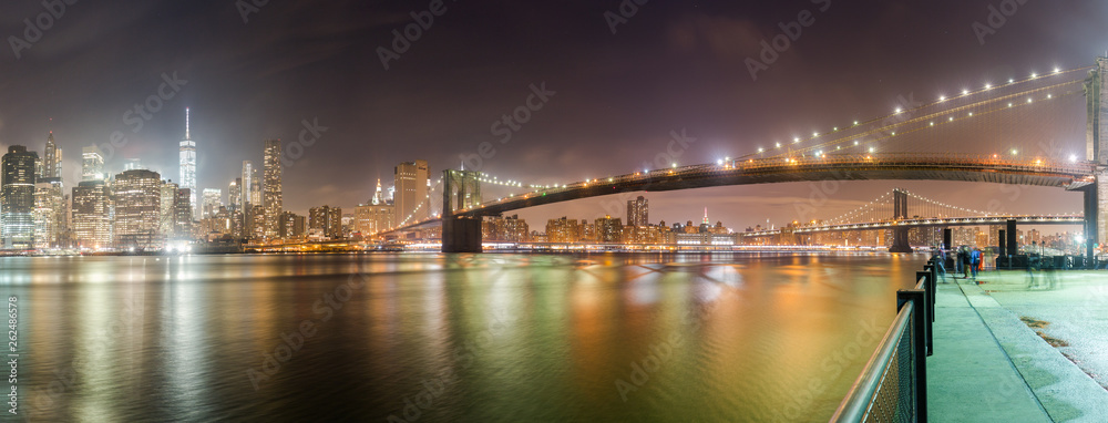A panorama of the Brooklyn Bridge at night with the New York City skyline in the background. The East River is in front and parts of the Manhattan Bridge can be seen.