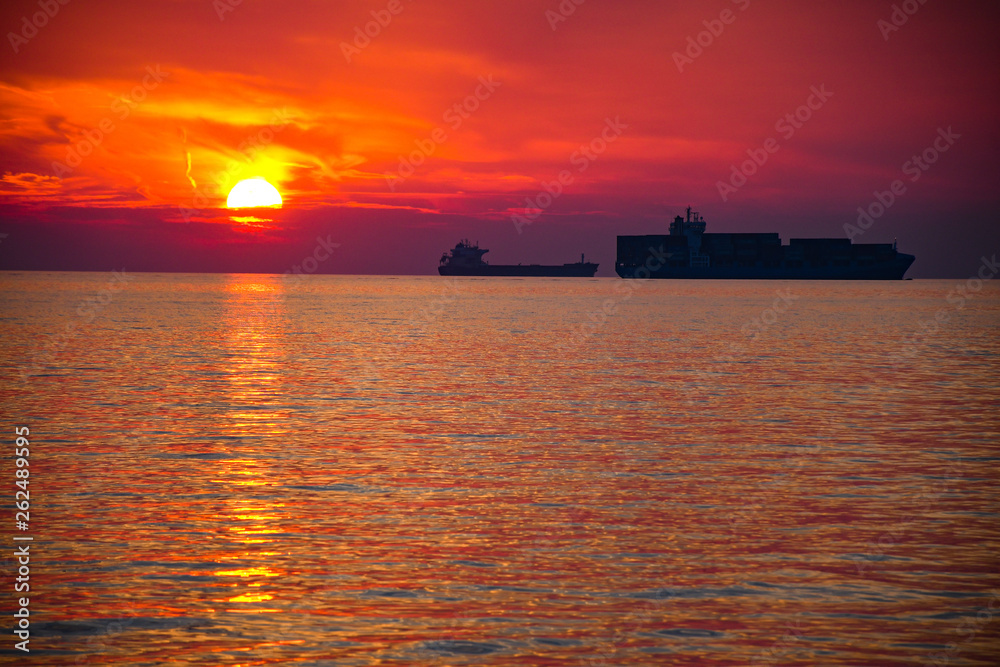 Small and big container tanker ship silhouette parked in a bay with a beautiful sunset sky and reflection in the ocean sea