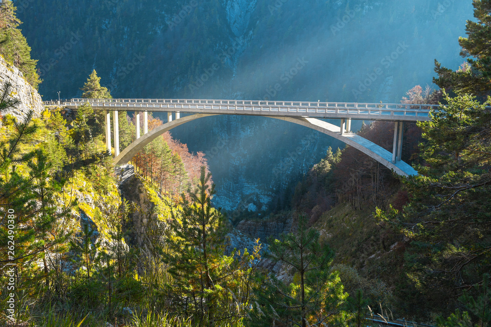 Fototapeta premium Bow shaped bridge over the river close to a road that is headed to Mangart saddle high in Julian alps on a sunny beautiful autumn day with colorful scenery and sun rays