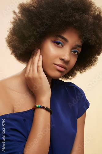 Portrait of African girl with blue eyeliner, wearing navy blue T-shirt with naked shoulder and versicolor stony bracelet. The girl is touching her neck, tilting her head against beige background.