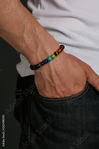 Cropped closeup shot of man's hand with stony bracelet, adorned with semiprecious stones. The guy in black jeans and white T-shirt is putting his hand into a pocket, posing on the dark background. photo