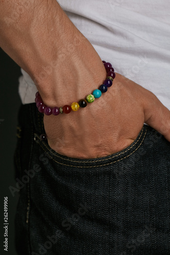 Cropped closeup shot of guy's hand with stony bracelet with different multicolor stones. The man in black jeans and white T-shirt is putting his hand into a pocket, posing on the dark background.