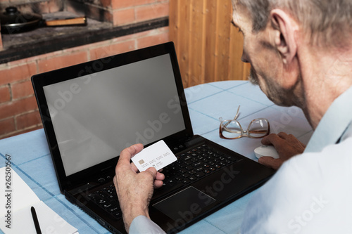 Senior man holding white credit card in front of laptop with blank screen for online shopping