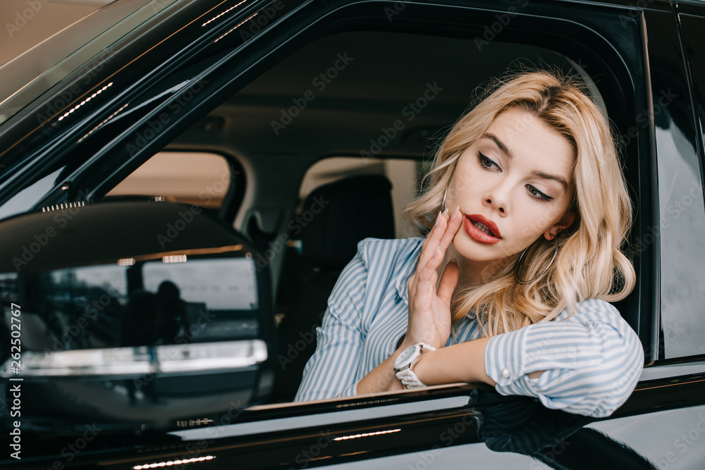 attractive blonde girl looking at car mirror while touching face