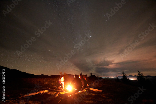 Three tourists travelers, two men and young woman sitting on big logs by burning campfire on grassy mountain valley with small spruce trees enjoying beautiful camping night under starry cloudy sky.