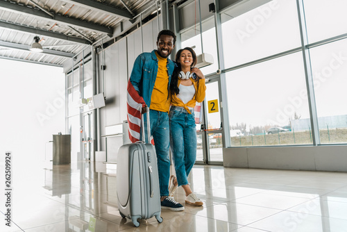 happy african american couple with american flag and luggage walking  in departure lounge in airport