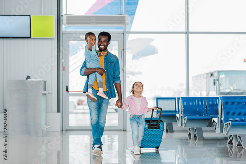 smiling african american father walking with happy children and baggage along waiting hall in airport