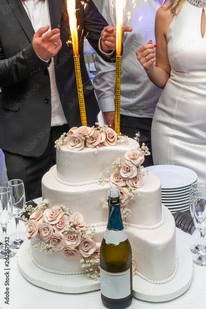 Three-tiered white wedding cake with sparklers and champagne on restaurant table ready for bride and groom