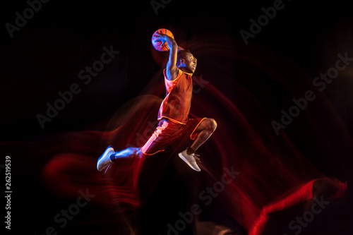 See the light of win. African-american young basketball player of red team in action and neon lights over dark studio background. Concept of sport, movement, energy and dynamic, healthy lifestyle.