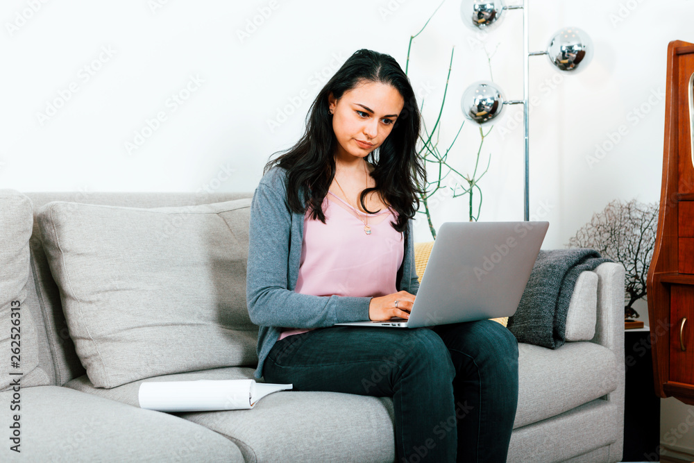 focused young woman sitting on sofa and using laptop - working and studying, home office concept