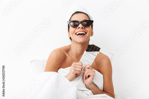 Woman with towel on head lies in bed under blanket isolated over white wall background wearing sunglasses eat chocolate.