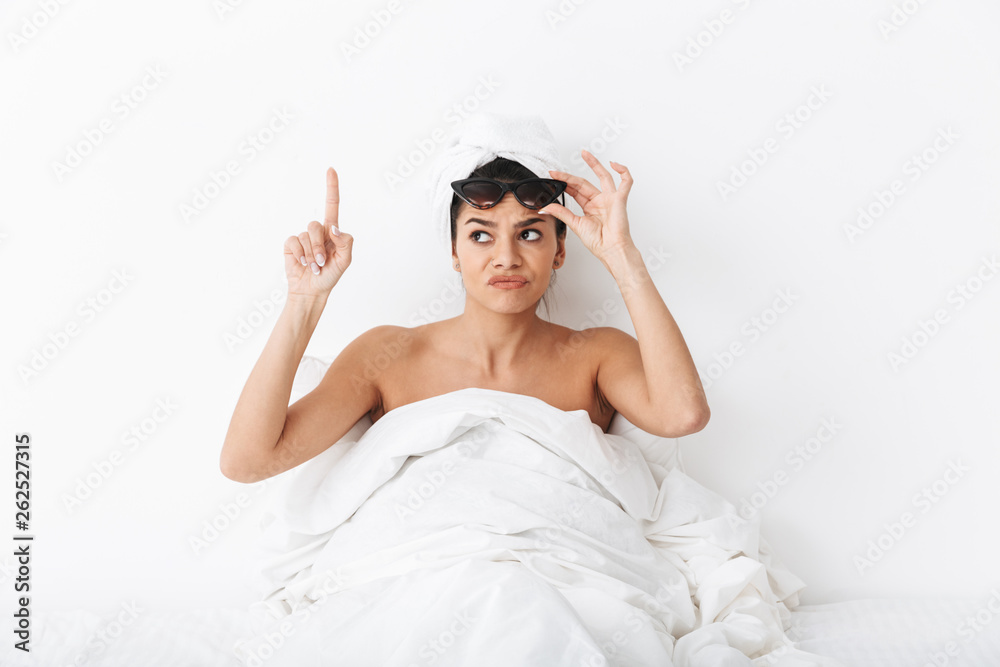 Woman with towel on head lies in bed covering body under blanket isolated over white wall background wearing sunglasses pointing. Woman with towel on head lies in bed covering body under blanket isolated over white wall background wearing sunglasses pointing.
