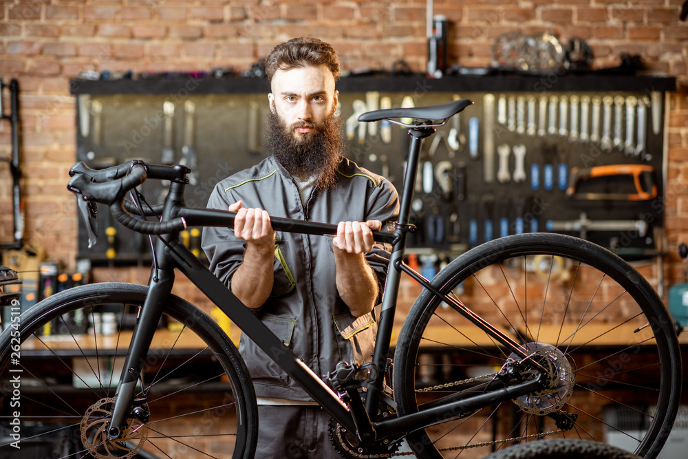 Obraz premium Portrait of a handsome cheerful repairman in workwear holding sports bicycle at the workshop