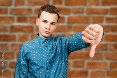 White young man dressed in blue shirt showing thumbs down on brick wall background photo