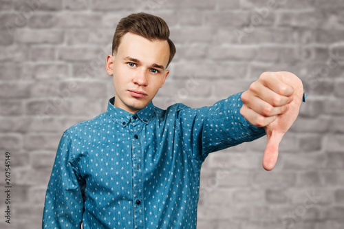 White young man dressed in blue shirt showing thumbs down on brick wall background photo