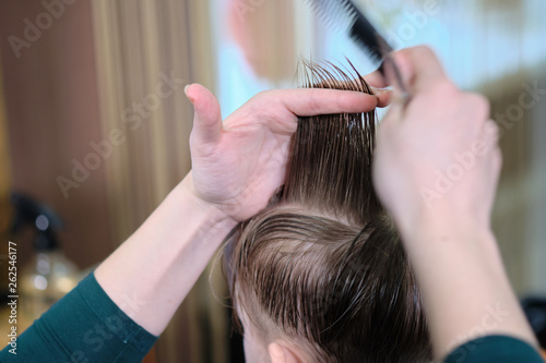 Barber shop. Hairdresser makes hairstyle to a boy with hair scissors and black comb. 