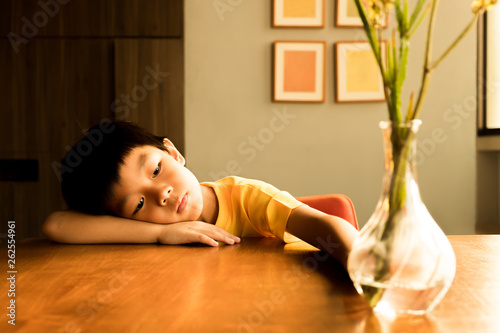 a little boy sitting on the wooden table under the sunlight in the evening with lonely feeling at home. he has no friends to play with him.