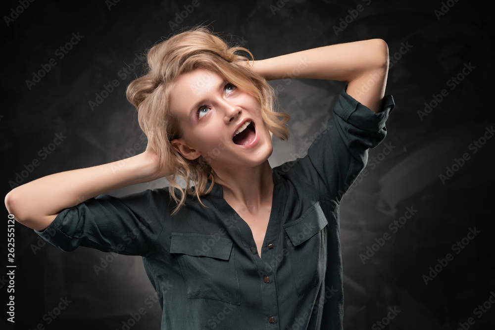 Beautiful young blonde girl with disheveled hairstyle and nude makeup, wearing a shirt and jeans emotionally posing on a gray background. She screams and holds on to her head
