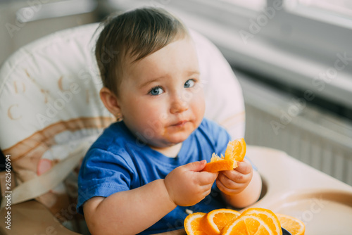 a little girl in a blue t-shirt and a blue plate sitting in a child s chair eating an orange