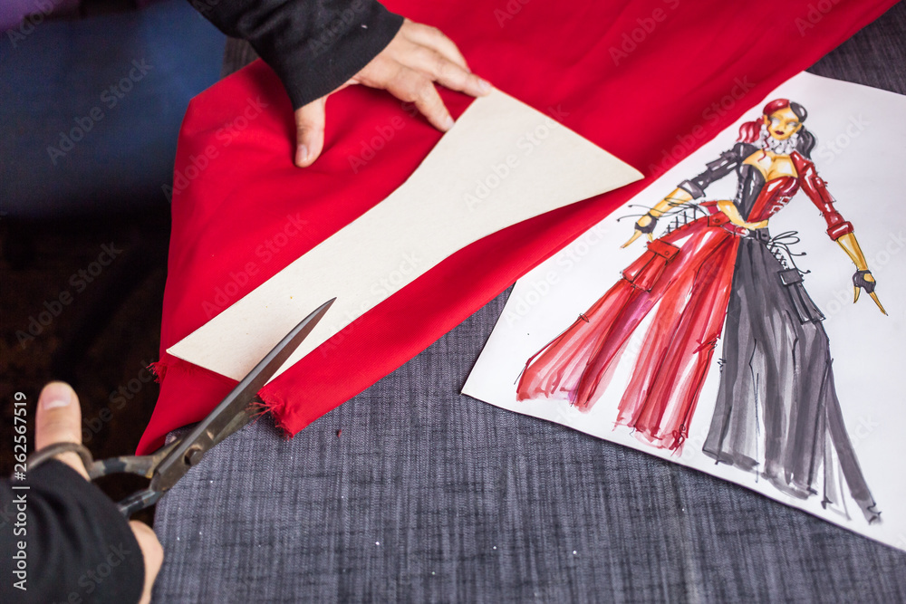 Fotka Close up photo of tailor s hand cutting with scissors piece of red fabric with the illustration of the dress next to it. Prom dress preparation. Fashion design concept ze slu by Stock
