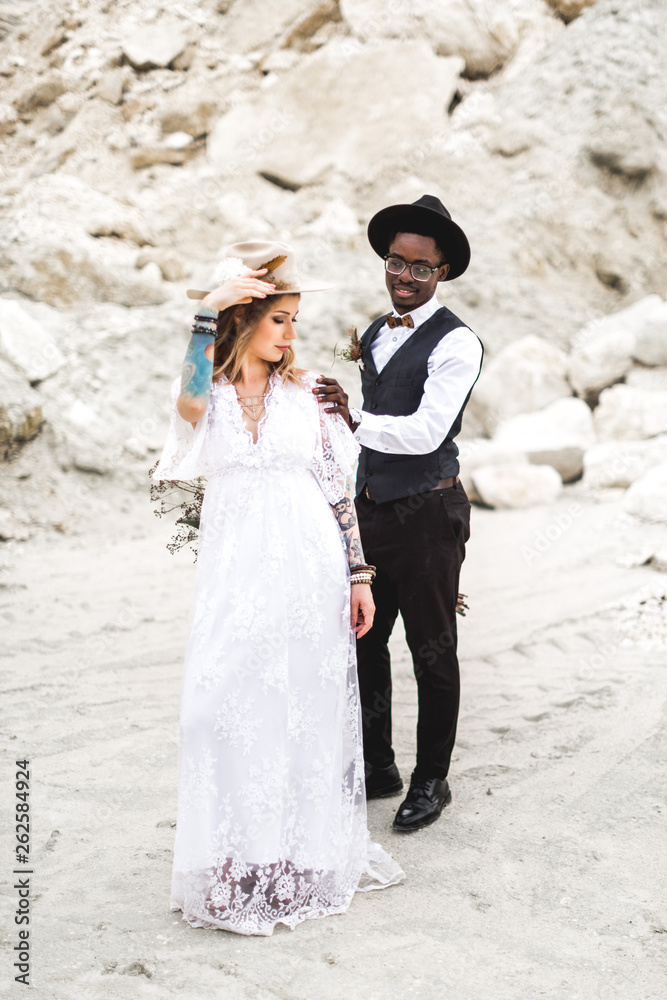 Handsome Afro American groom in stylish blask suit and hat walking in desert sand canyon and touching his pretty bride in long beautiful boho wedding dress and stylish hat. Canyon, desert love story