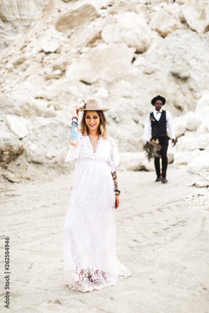 A beautiful bride in boho rustic dress is standing in a canyon wearing a stylish hat. Stylish African American groom in black suit standing on the background. Wedding in desert, canyon, love story