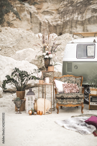 Green hippy bus, vintage armchair with pillows, retro radio, golden metal decorations with plants, succulents, flowers and candles, fur carpet on the background of desert landscape. Boho decor