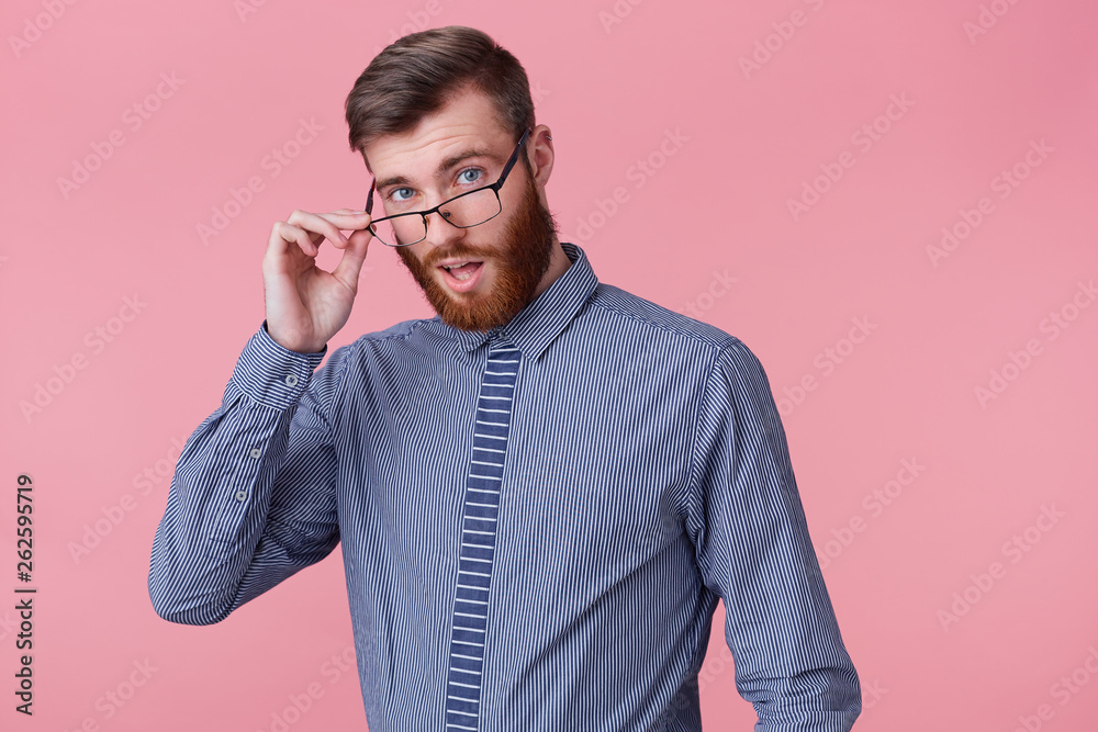 Close up of young successful man looks to the camera through his glasses isolated over pink background, open mouth, a little surprised.
