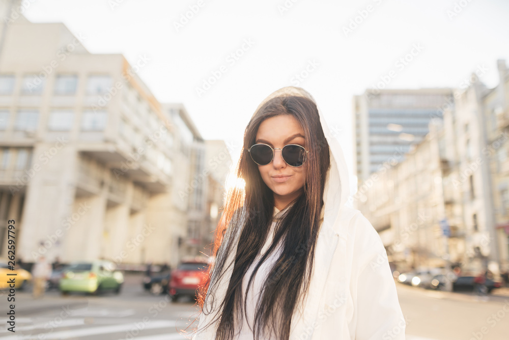 Street portrait of an attractive girl wears sunglasses and a white jacket, stands on the background of a city landscape at sunset, looks at the camera and smiles. Fashionable Urban Girl Photo.