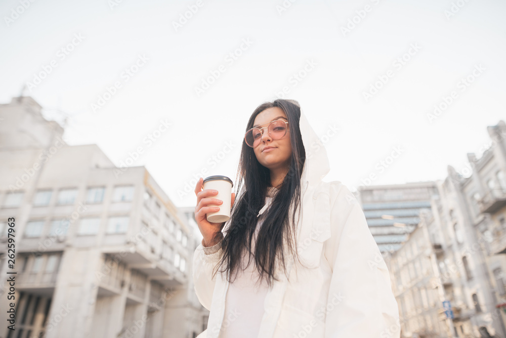 Street portrait of a stylish girl in casual clothing,standing on the street with a cup of coffee in his hand, looks camera.Woman in a stylish white jacket against the backdrop of the city architecture