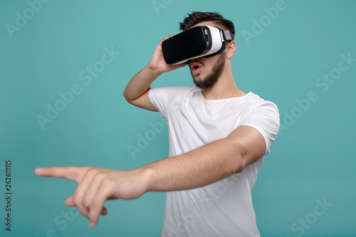 Attractive bearded man trying VR headset and poniting finger. Young man exploring another world with virtual reality goggles on the colorful background.