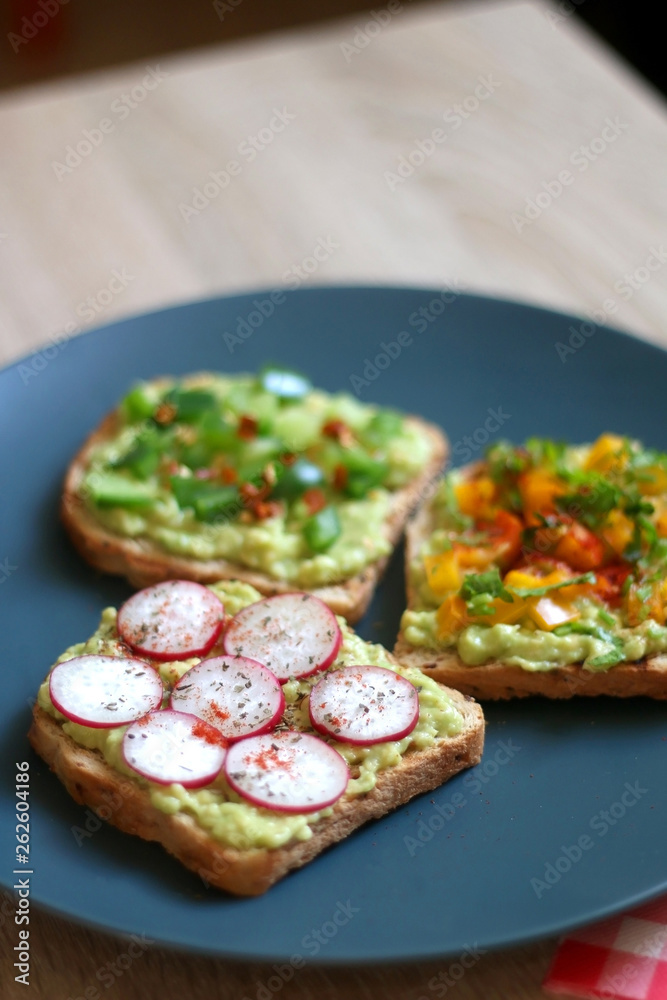Three slices of toast with mashed avocado and various vegetable and herb toppings. Selective focus. Three slices of toast with mashed avocado and various vegetable and herb toppings. Selective focus.