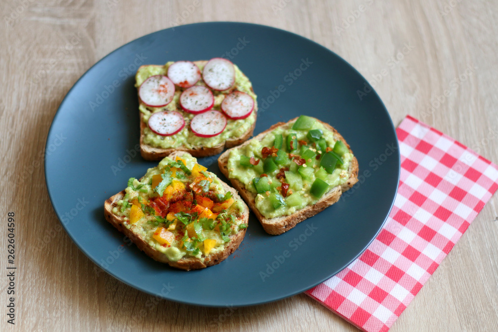 Three slices of toast with mashed avocado and various vegetable and herb toppings. Selective focus. Three slices of toast with mashed avocado and various vegetable and herb toppings. Selective focus.