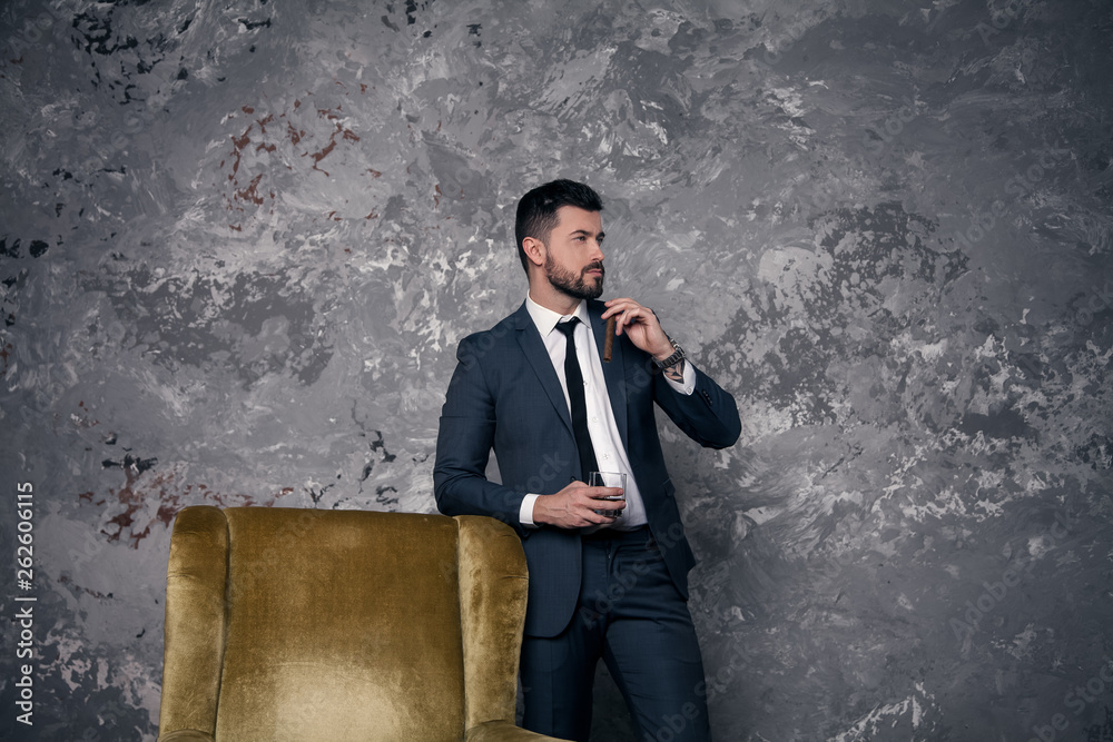 Good looking handsome businessman is taking a rest in the office standing by the chair and drinking whisky with a cigar and thinking. wearing suit and a tie Good looking handsome businessman is taking a rest in the office standing by the chair and drinking whisky with a cigar and thinking. wearing suit and a tie