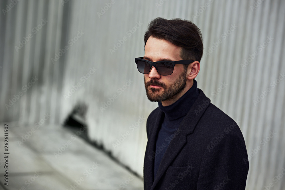 Obraz premium Fashion beard style business handsome male model in fashion sunglasses and blue coat with serious concentrated look posing in style clothing on wall outdoors background.