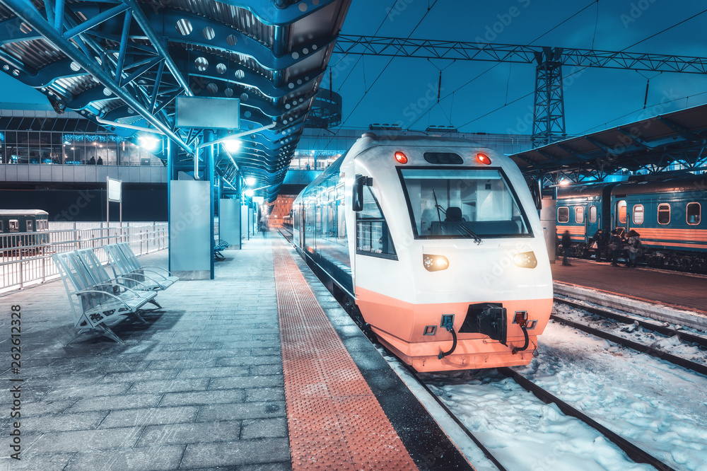 High speed train on the railway station at night in winter. Urban landscape with modern commuter white train on the railway platform with illumination at dusk. Intercity. Passenger railroad travel