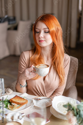 Nice redhead woman eat breakfast in cafe. Portrait of young charming female drinking coffee and toasted sandwich in modern coffee shop.