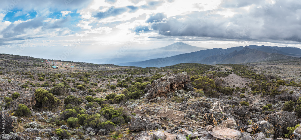 Panoramic view of the Shira Cave Camp site on the Machame hiking route on Mt Kilimanjaro, Tanzania. Mount Meru is behind sun rays in the background under a dramatic cloudy sky, with rocks and foliage.