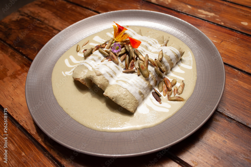 Platillo mexicano, enchiladas en salsa verde de pipián decoradas con pepitas y flores sobre platillo de cerámica y mesa de madera Platillo mexicano, enchiladas en salsa verde de pipián decoradas con pepitas y flores sobre platillo de cerámica y mesa de madera