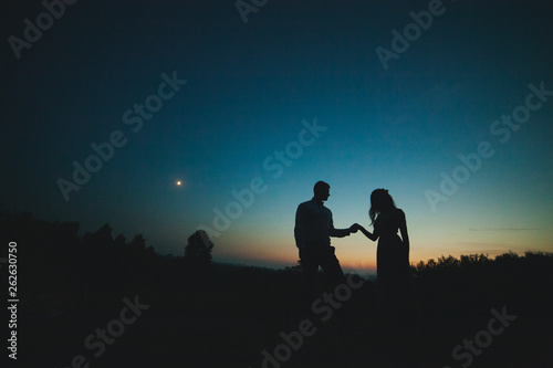 Silhouettes of a young couple on a sunset background  the moon in the sky. The bride and groom stand against the setting sun. The guy and the girl against the sky at sunset. The concept of love.