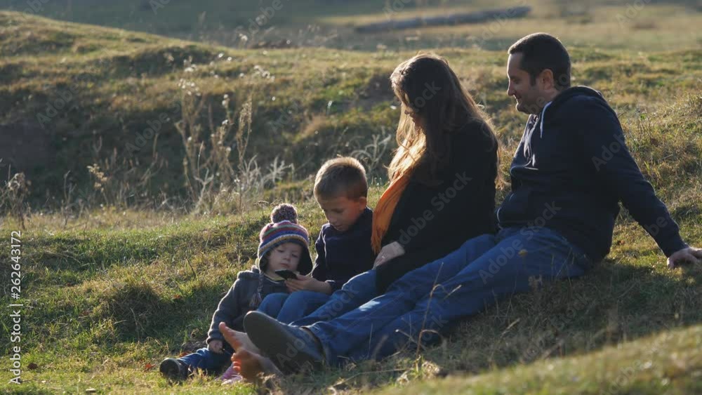 Family portrait, parents sitting on grass and children looking at mobile phone in nature Family portrait, parents sitting on grass and children looking at mobile phone in nature