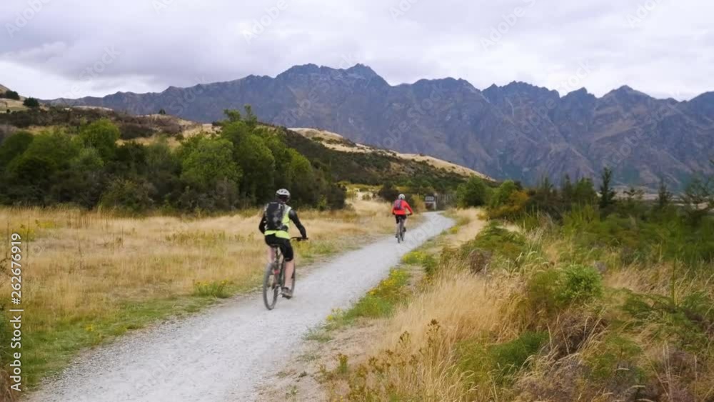 couple of mountain bikers riding along gravel track below Remarkables mountain range in Queenstown, New Zealand couple of mountain bikers riding along gravel track below Remarkables mountain range in Queenstown, New Zealand