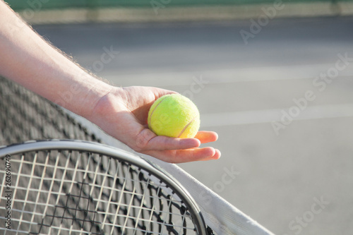 Sportsman is playing tennis on court outdoor. Male hand holds tennis racket and gives yellow ball over net. Man has workout. Sports activities in summer.