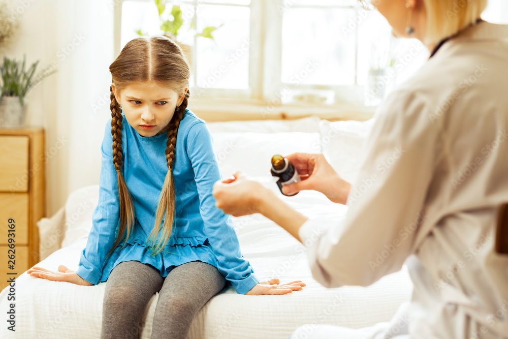 Small sick girl dissatisfiedly looking at pediatrician pouring throat syrup Small sick girl dissatisfiedly looking at pediatrician pouring throat syrup