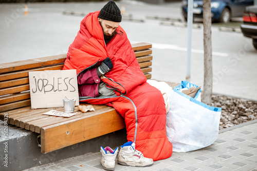 Homeless and jobless beggar sitting on the bench wrapped with sleeping bag begging money near the business center